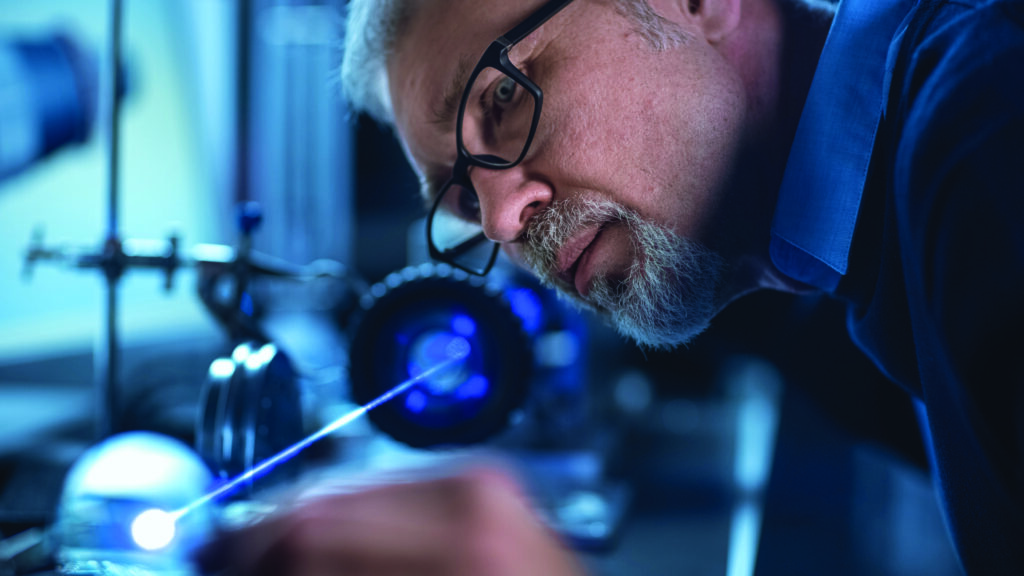 Close-up Portrait of Focused Middle Aged Engineer in Glasses Working with High Precision Laser Equipment, Using Lenses and Testing Optics for Accuracy Required Electronics